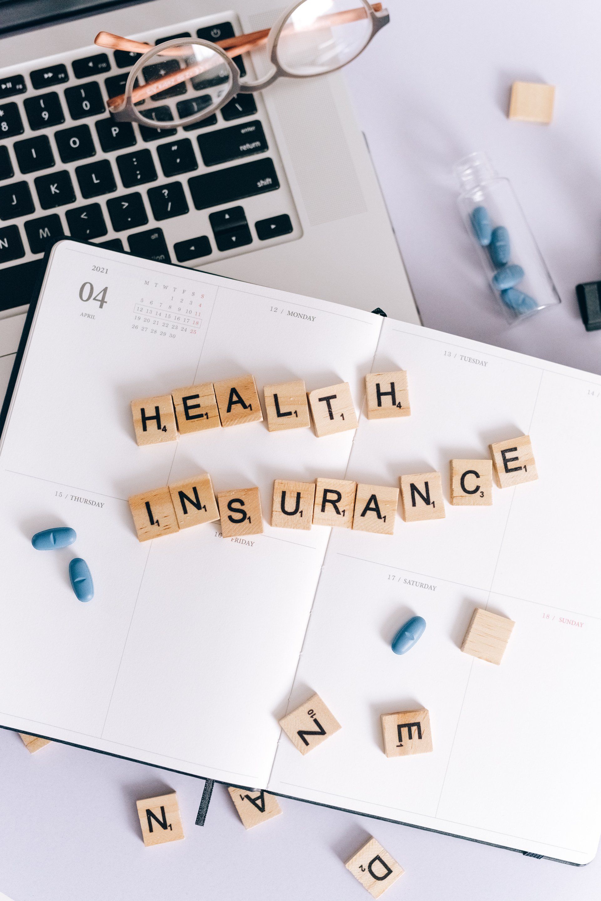 Health insurance spelled with letter tiles on a notebook beside a laptop and glasses