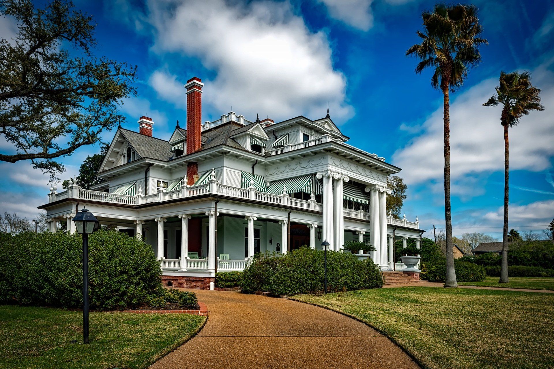 Historic white Victorian mansion with wraparound porch and red chimney, set on a lawn with palm trees and blue sky