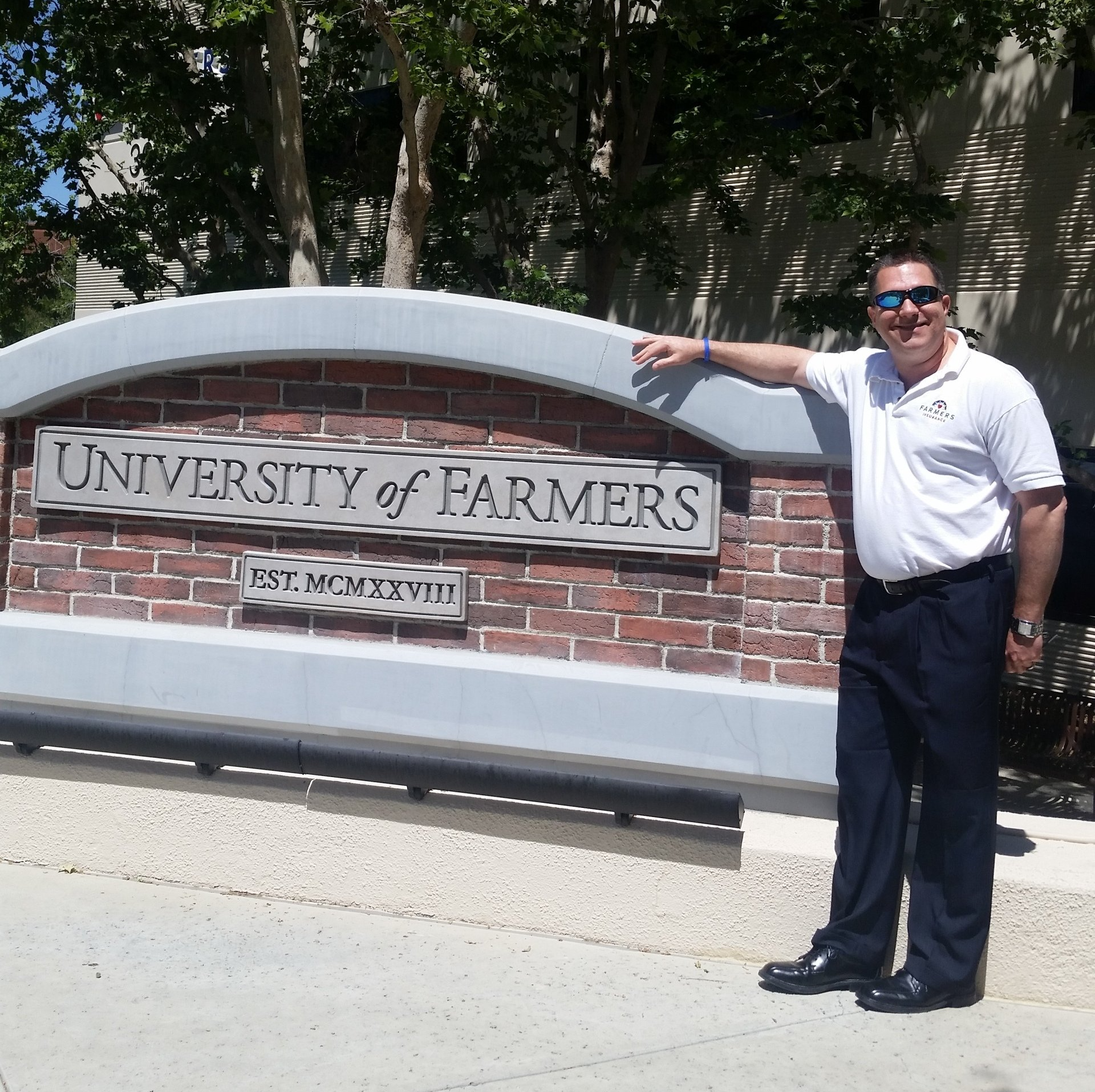Man standing beside a brick “University of Farmers” sign, pointing at it outdoors.