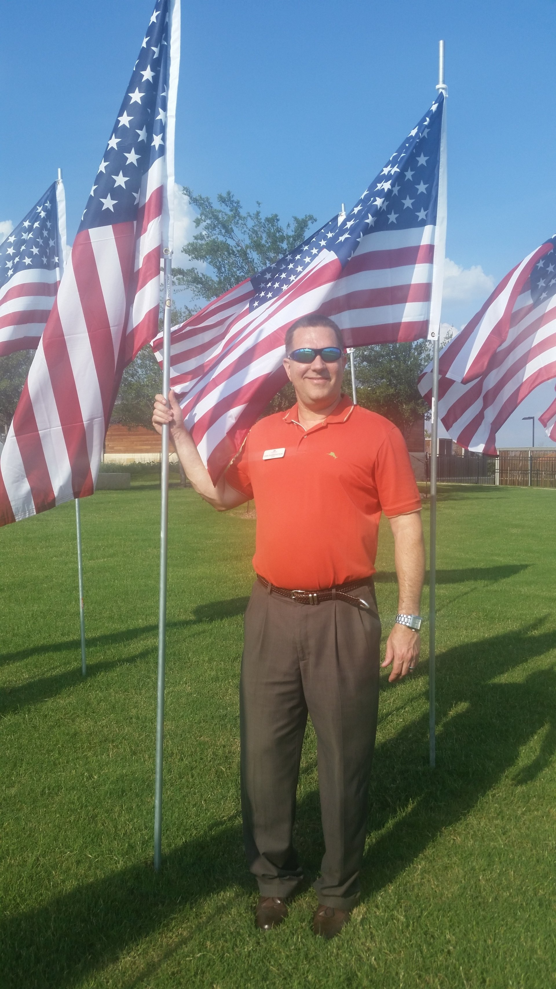Man in red shirt standing on grass among large American flags in a sunny field